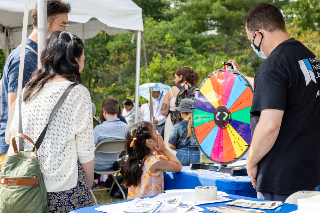 A young girl in a floral dress excitedly watches a colorful spinning prize wheel at the Telling Tales Festival, Canada’s leading children’s literacy festival. A staff member from the Hamilton Public Library operates the wheel at an activity booth, engaging families in a fun, interactive experience. A woman with a green backpack and a man stand nearby observing. In the background, festival attendees, including families and children, enjoy various literary-themed activities under tents in a lush outdoor setting. Telling Tales is a non-profit organization dedicated to creating outstanding literary events and inspiring a love for reading.
