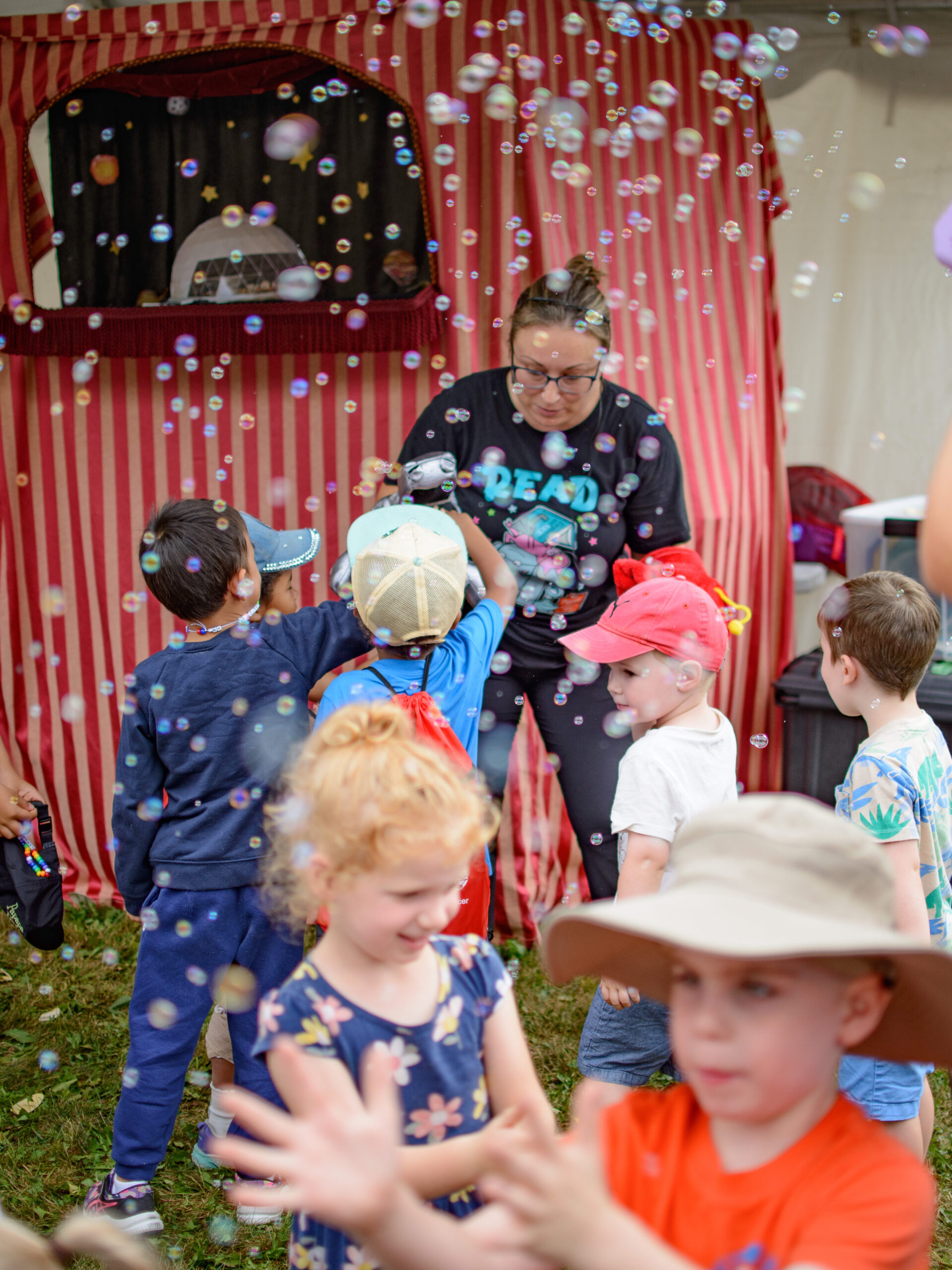 Children joyfully play with bubbles in front of a striped puppet show tent at the Telling Tales Festival, Canada’s leading children’s literacy festival. A performer in a black t-shirt interacts with the kids, creating an engaging storytelling experience. A woman in the foreground holds a bubble machine, adding to the magical atmosphere. The scene is filled with excitement as children laugh, reach for bubbles, and participate in the festival’s interactive programming. Telling Tales is a non-profit organization dedicated to inspiring a love for reading through outstanding literary events.